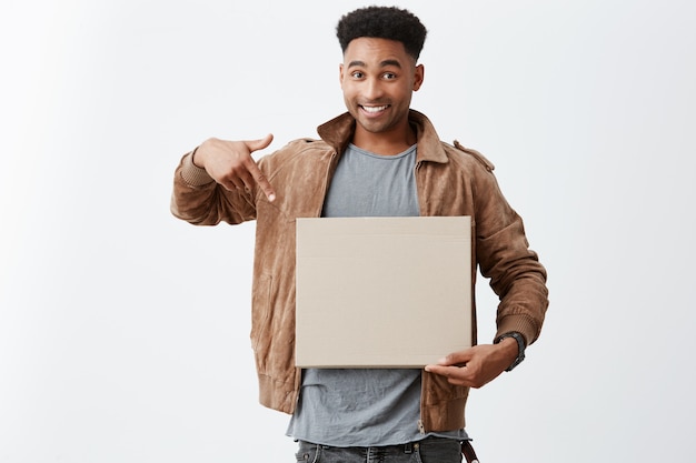 Take a look at this. Close up portrait of young beautiful black-skinned male university student with afro hairstyle in stylish autumn clothing pointing at carton board in hands with happy expression