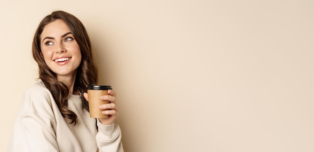 Takeaway and cafe concept beautiful feminine woman smiling holding cup of coffee posing against beig