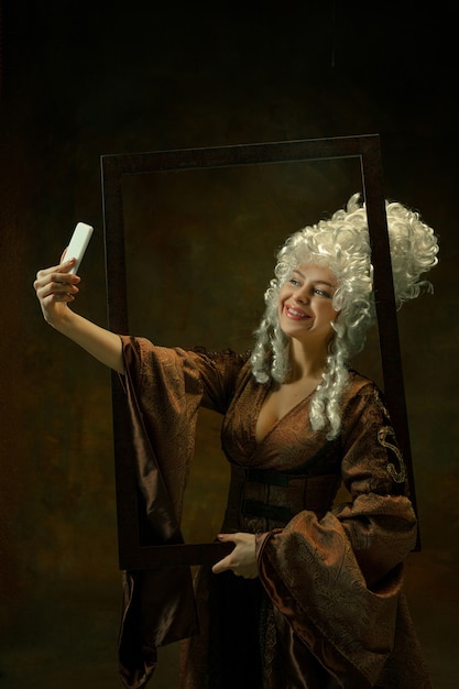 Free photo taking selfie. portrait of medieval young woman in vintage clothing with wooden frame on dark background. female model as a duchess, royal person. concept of comparison of eras, fashion, beauty.