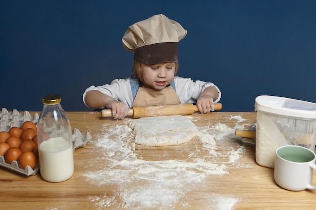 Talented charming little girl in chef hat and apron using rolling pin while kneading dough for homemade pizza.pasty. Childhood concept