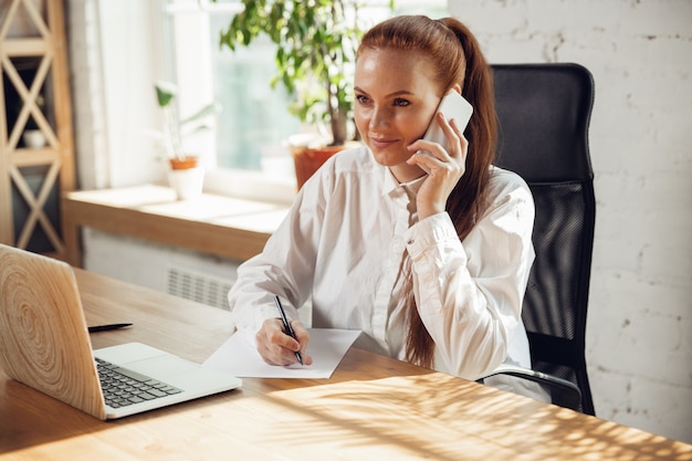 Free photo talking on phone. caucasian young woman in business attire working in office. young businesswoman, manager doing tasks with smartphone, laptop, tablet has online conference. concept of finance, job.