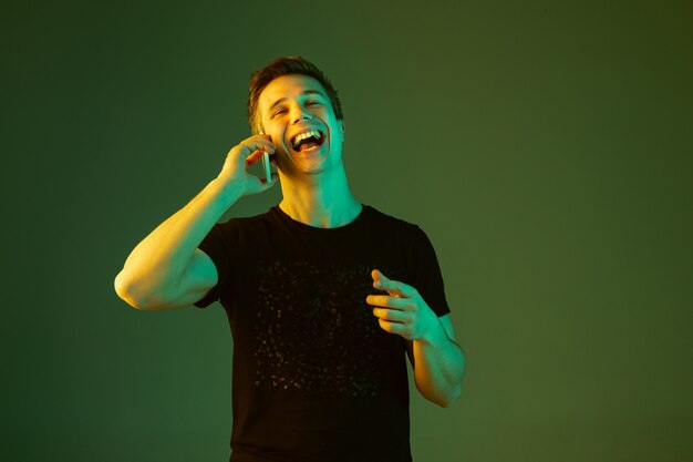 Talking on phone, laughting. Caucasian man's portrait isolated on green studio background in neon light. Beautiful male model in black shirt. Concept of human emotions, facial expression, sales, ad.