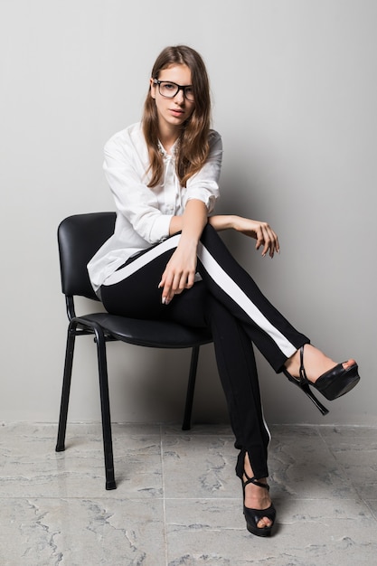 Free photo tall brunette girl in glasses dressed up in white t-shirt and black pants sits on office chair in front of white background