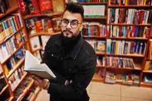 Free photo tall smart arab student man wear on black jeans jacket and eyeglasses at library with book at hands