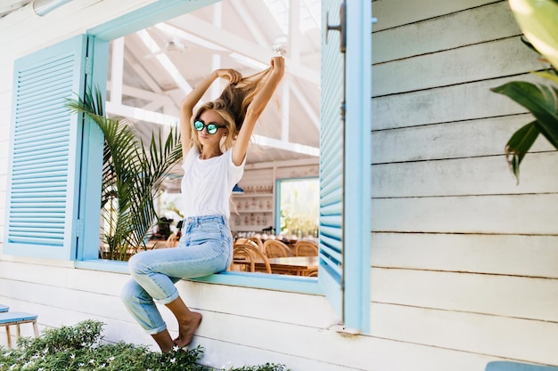 Free photo tanned barefooted woman in vintage jeans playing with her beautiful long hair photo of fashionable girl in sparkle sunglasses chilling on window sill