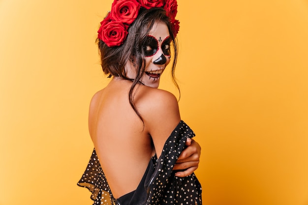 Tanned dark-haired girl happily poses in dress with open back. Lady with Halloween make-up surprised looking at camera.