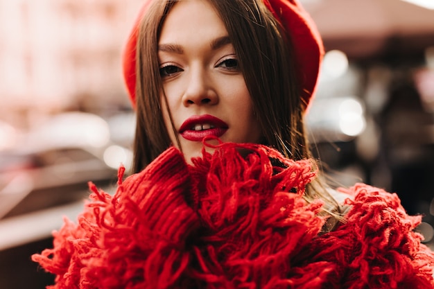 Free photo tanned dark-haired woman in red wool coat and beret looking at camera against background of city street.