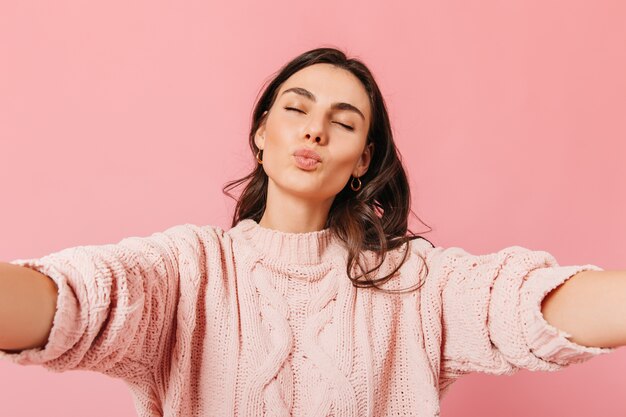 Tanned European girl with closed eyes sends kiss to camera. Lady in pink sweater wants hugs.