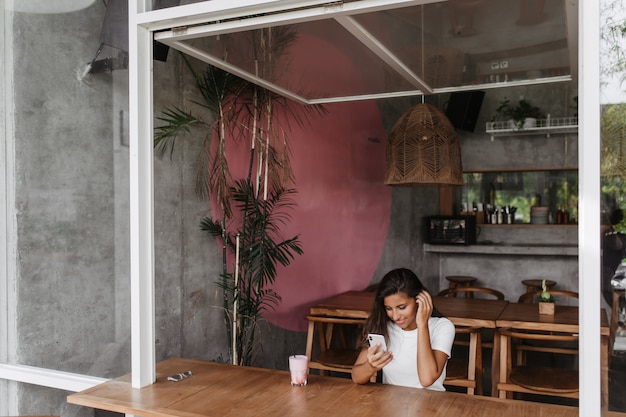 Free photo tanned lady is sitting in cafe with wooden furniture and watching video on phone