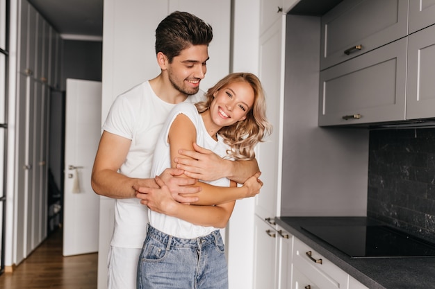 Tanned smiling man dancing with wife. Indoor portrait of couple embracing in cozy apartment.