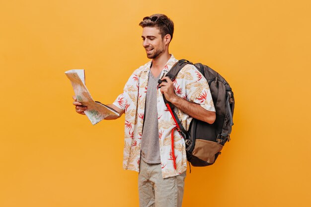 Tanned tourist in fashionable light cool outfit and sunglasses posing with backpack and map on isolated orange wall