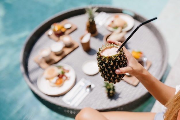 Free photo tanned woman holding sweet pineapple cocktail. female model posing during lunch in pool.