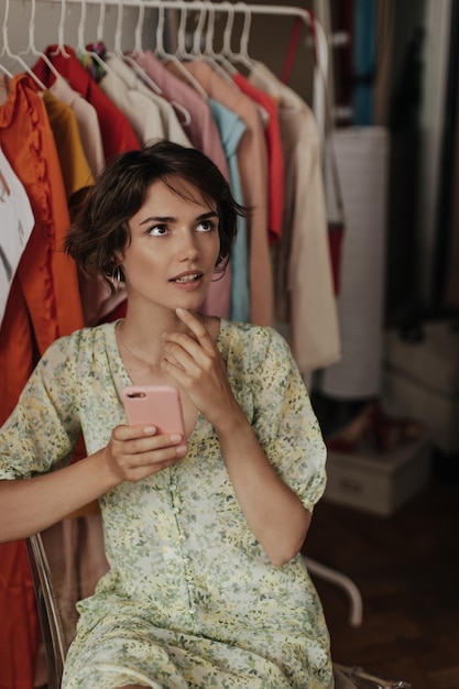 Free Photo tanned young attractive woman in green floral dress looks up holds phone in pink case and sits on chair in cozy dressing room