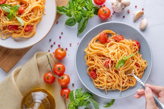 Tasty appetizing classic italian spaghetti pasta with tomato sauce, cheese parmesan and basil on plate and ingredients for cooking pasta on white marble table. Flat lay top view copy space.