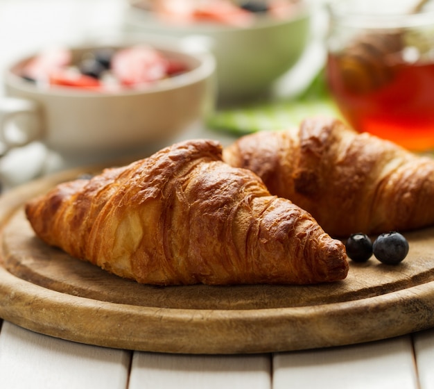 Tasty beautiful croissants on wooden board. Traditional continental breakfast. Granola with fruits and honey on background.