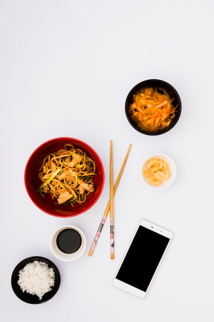 Free photo tasty noodles in bowl near salad; ginger pickle; soya sauce and steamed rice with mobile phone on white background