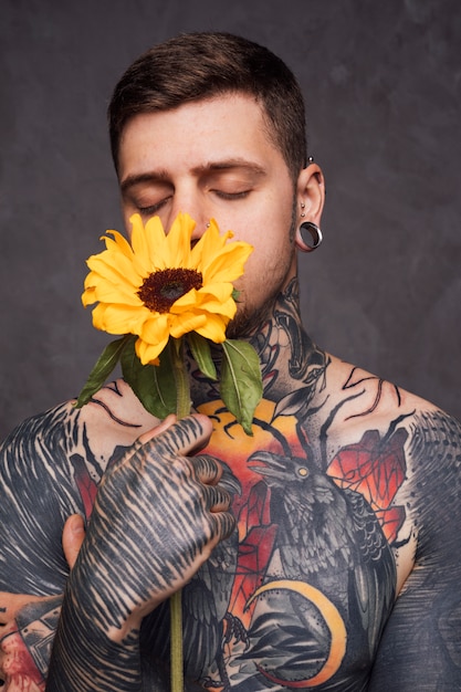 Free photo tattoo young man with holding sunflower in front of his mouth against grey background