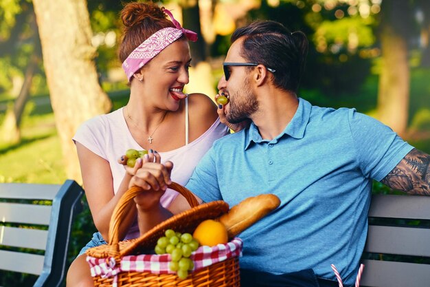 Tattooed bearded male and redhead female are having a picnic on a bench in a park.