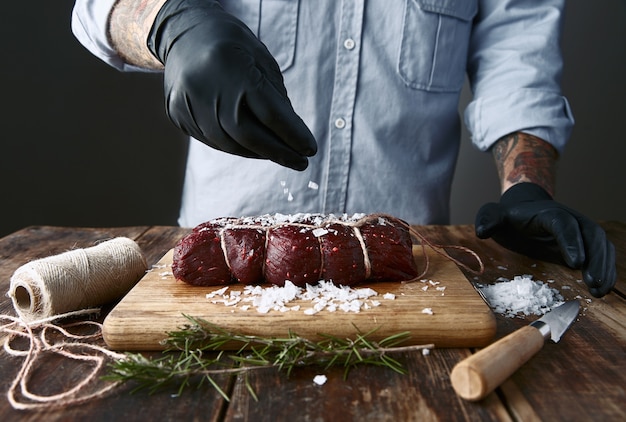 Free photo tattooed butcher in black gloves salts tied piece of meat to smoke it.