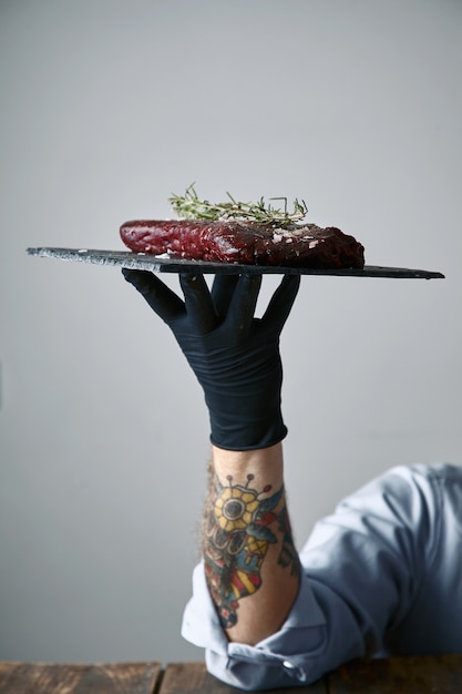 Free Photo tattooed hand in black glove holds stone plate with steak ready to cook