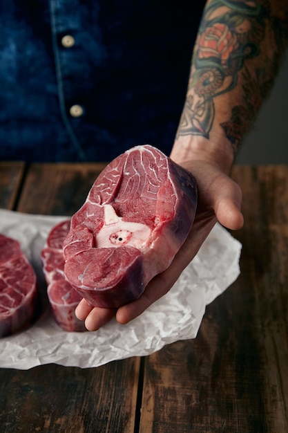 tattooed hand offers a piece of meat stake above two steaks on craft paper, close up