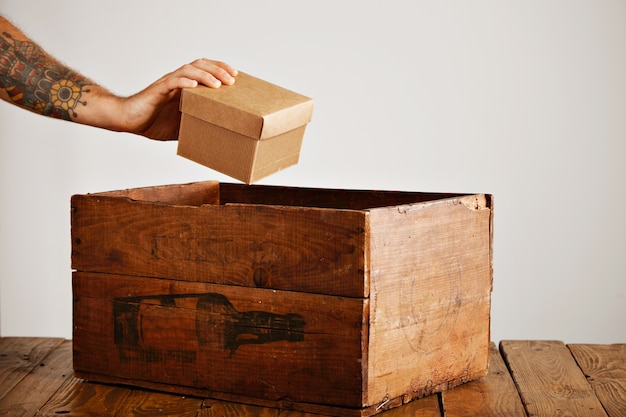 Free photo tattooed hand picks up cardboard package from old wooden crate on rustic table, isolated on white