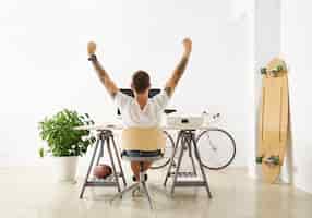 Free photo tattooed lucky freelancer in front of his working space, surrounded with his hobby toys longboard, vintage bicycle and green plant, stretching his hand in air while making break