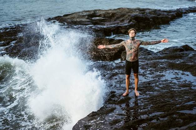 A tattooed man lies on the edge of a cliff. splashes of ocean waves.