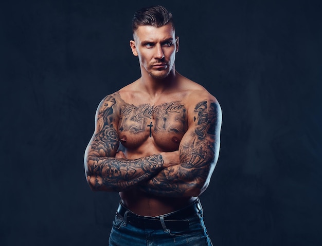 A tattooed muscular shirtless man with stylish hair posing at the camera over dark background.