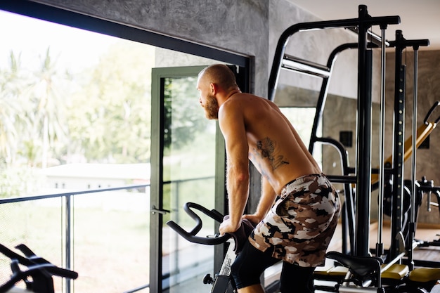 Free Photo tattooed muscular strong bearded man work out cardio on bicycle in gym near big window with view on trees outside