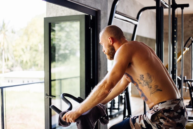 Free Photo tattooed muscular strong bearded man work out cardio on bicycle in gym near big window with view on trees outside