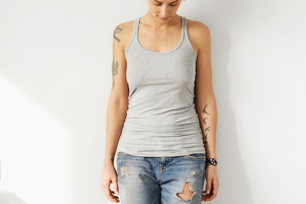 tattooed young European female model looking down at grey blank A-shirt. Cropped portrait of teenge woman dressed in ripped jeans and tank top posing indoors. Youth and fashion