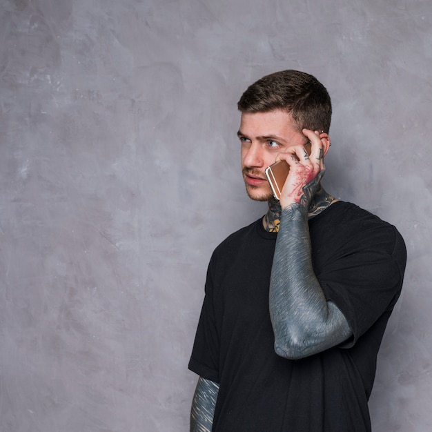 Tattooed young man talking on mobile phone against textured gray background