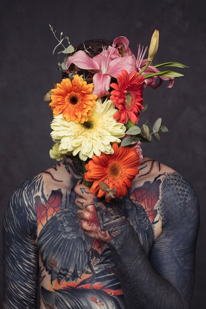 Tattooed young man with pierced ear and nose holding flower bouquet in front of his face