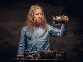 Free photo tea ceremony concept. portrait of a redhead hipster male with long hair and full beard dressed in a blue shirt, concentrates on making tea, using a handmade tea set. isolated on a dark textured backgr