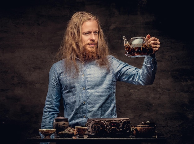 Tea ceremony concept. Portrait of a redhead hipster male with long hair and full beard dressed in a blue shirt, concentrates on making tea, using a handmade tea set. Isolated on a dark textured backgr