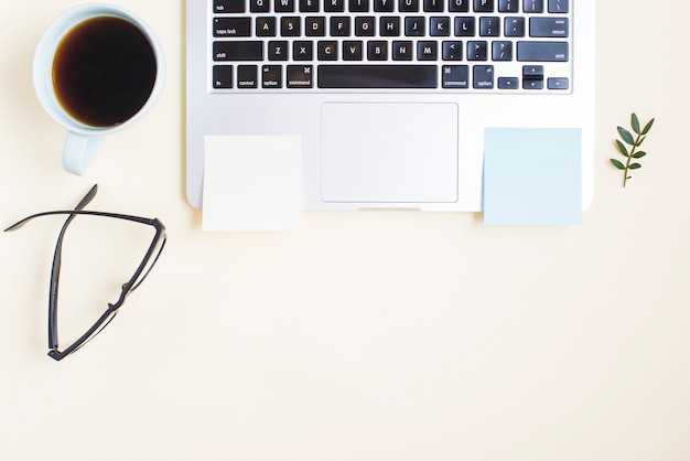 Tea cup; eyeglasses and blank adhesive notepad on laptop against beige backdrop