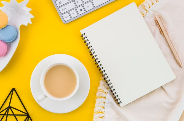 Tea cup; macaroons; spiral notebook; pen on tablecloth against yellow background