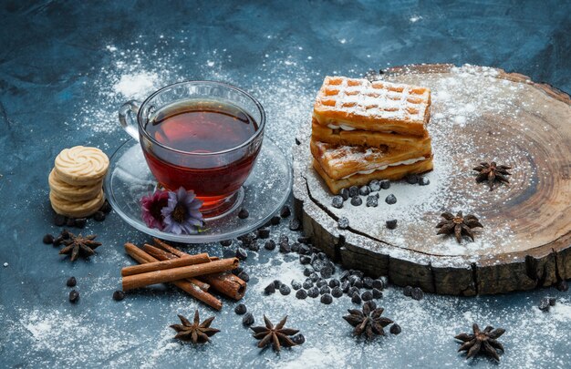 Tea in a cup with waffle, biscuits, choco chips, spices high angle view on blue grunge and wooden board