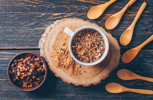 Tea herbs in a bowl and cup with spoons top view on a wood stub and dark wooden background