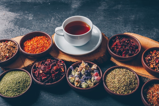 Tea herbs in a bowls with wood stubs and a cup of tea high angle view on a dark textured background. space for text