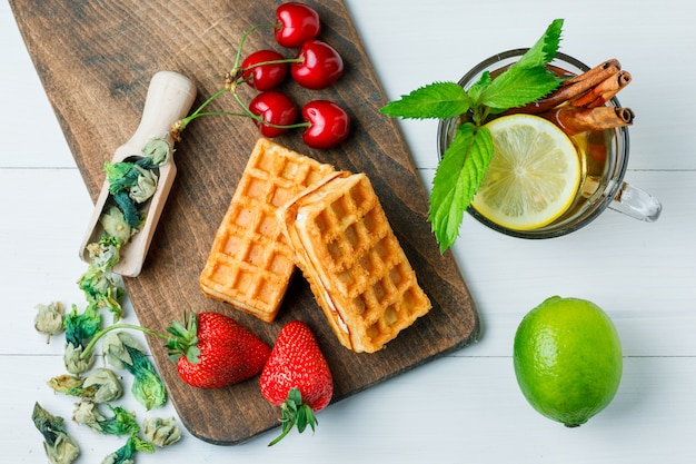 Free photo tea with lemon, waffle, dried herbs, fruits, mint, cinnamon in a cup on wooden and cutting board, flat lay.