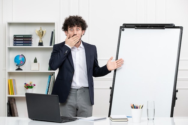 Teacher brunette instructor with computer in suit and whiteboard classroom covering mouth shocked