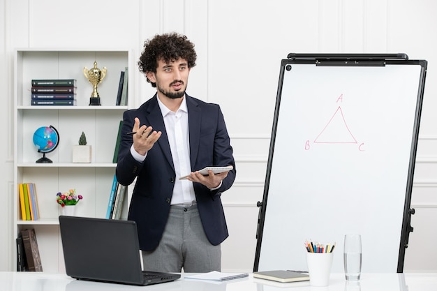 Teacher brunette instructor with computer in suit and whiteboard in classroom looking seriously