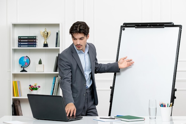 Teacher smart instructor in grey suit in classroom with computer and whiteboard talking to student