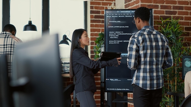 Team of coders analyzing code on wall screen tv comparing errors using digital tablet next to programer working with neural network. Software engineers collaborating on coding group project.