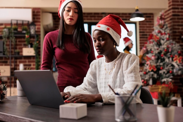 Team of coworkers working on business in workplace decorated with christmas lights, tree and holiday ornaments. Using laptop in festive company office with seasonal xmas decorations.