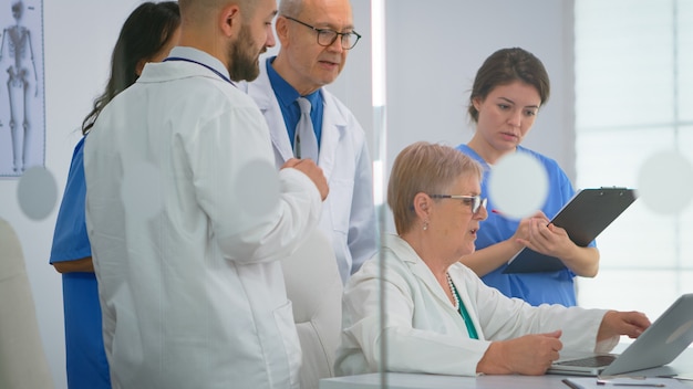 Team of doctors standing in conference hospital room, senior doctor discussing about treatment for patient looking in laptop. Coworkers in white coats working together analysing symptoms of disease