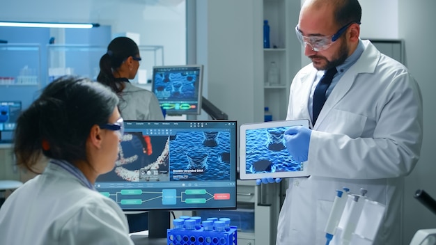 Team of industrial scientists, engineers, developers innovating new vaccine, doctor pointing on tablet explaining virus evolution to coworker