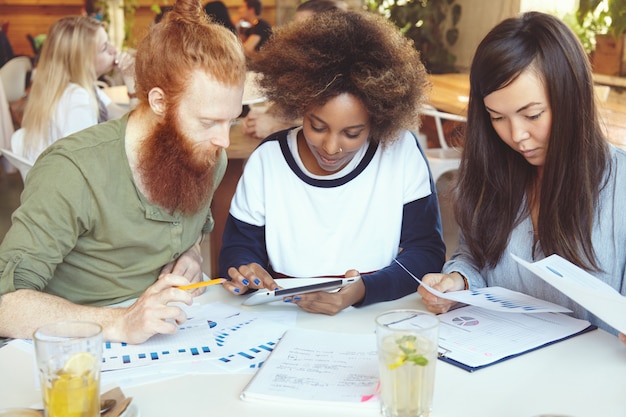 Free photo team of marketing experts developing business strategy at cafe african woman presenting business plan to her partner with red beard on digital tablet while their asian colleague analysing graphs